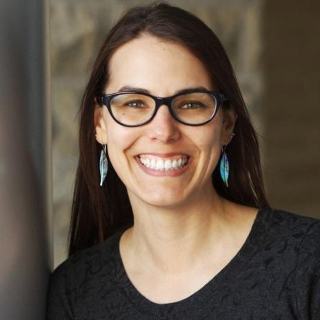 a woman with long hair and feather earrings smiles for a portrait against a stone wall