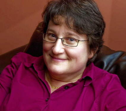 Dr. Lynne Mitchell sits in a chair wearing glasses and a maroon shirt.