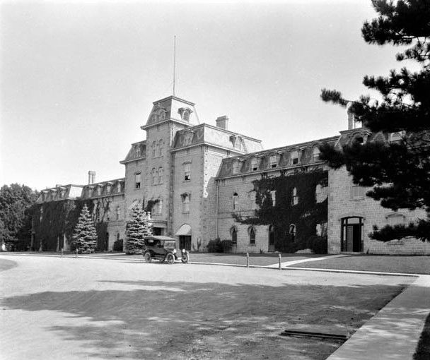 Moreton Lodge at the Ontario Agricultural College. An old car is parked outside a long, rectangular-shaped building with three central taller units, an antenna in the middle.