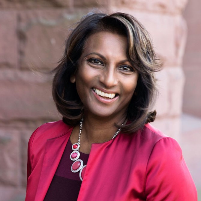 Indira Naidoo-Harris leans against a pale, red brick wall.