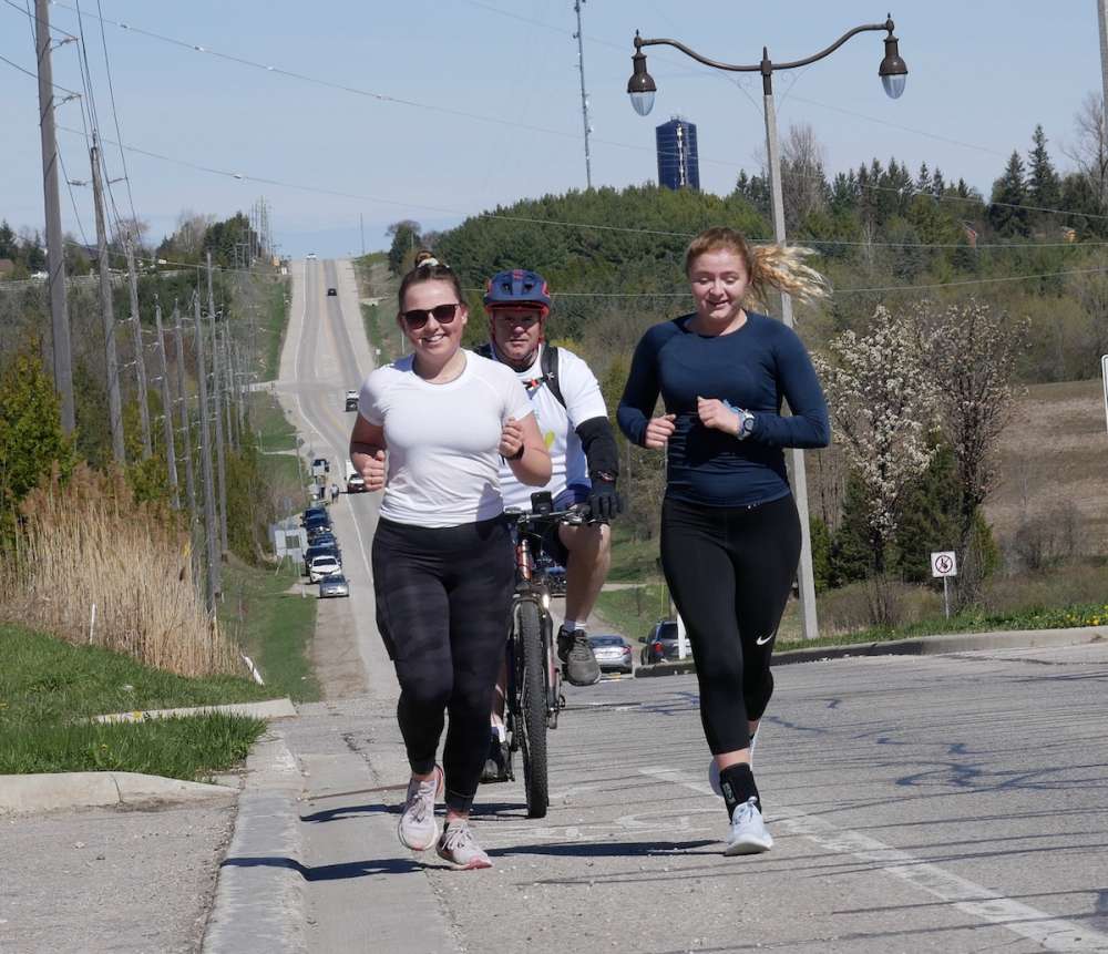 Two young women running along a highway, accompanied by man on bike.