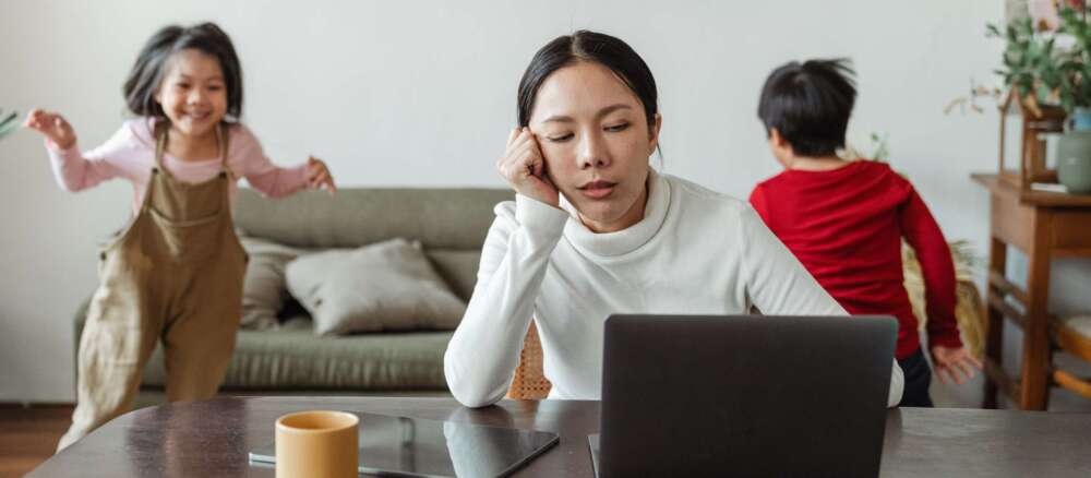 A tired-looking mother sits at a laptop while two children run in circles behind her.