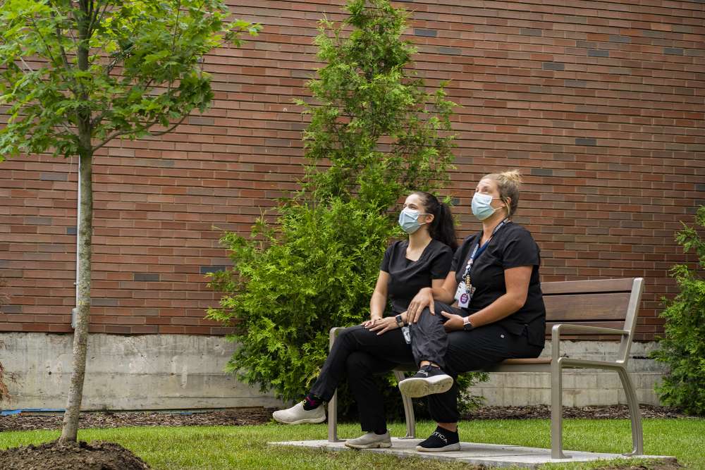 Two hospital staff at St. Peter’s Hospital in Hamilton sit on a bench and look at a tree planted as part of the One Bench, One Tree project.