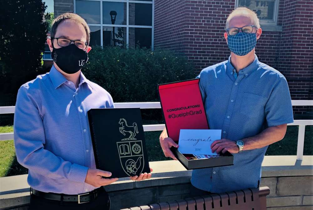 Brett Shepherd, director of Ridgetown campus with Dr. Simon Lachance, assistant dean of academic at Ridgetown campus, from the waist up. They are holding black and red boxes with the U of G logo on them. These are graduate boxes sent to all the graduates. There is a building the background.