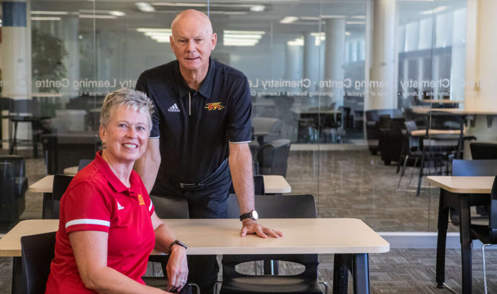 Joy and Larry Pearson in U of G's McLaughlin Library. Joy sits in a chair next to a table. Larry stands behind her and leans on the table. Both wear Gryphon shirts