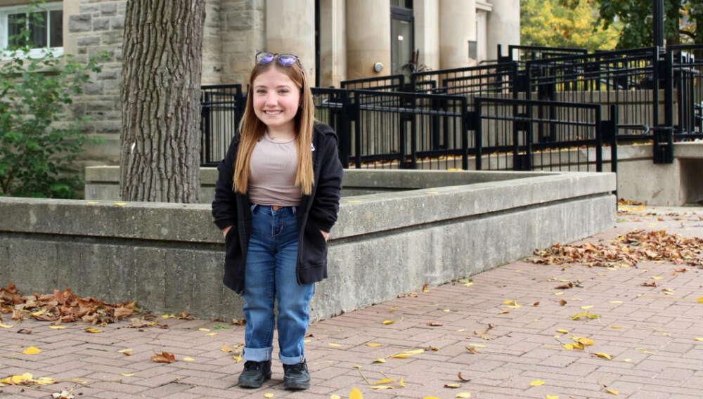 A woman of short stature smiles for the camera on the leaf-strewn U of G campus
