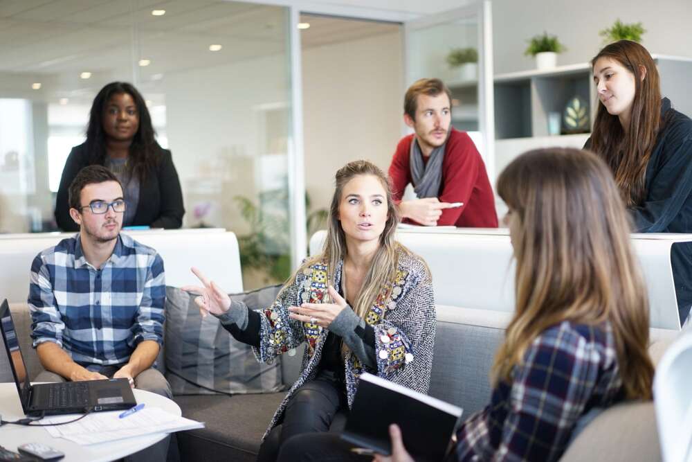 Several people stand around a workplace sofa in discussion