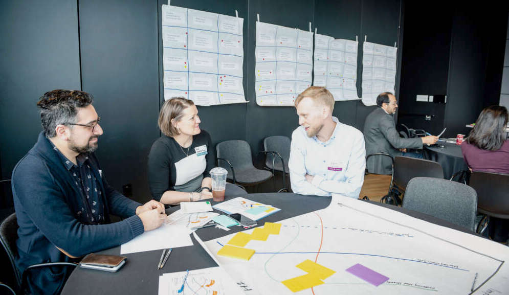 Three people chatting around a round table shattered with papers and notes