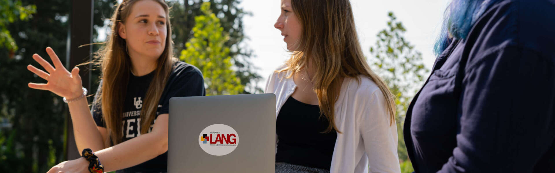 three female students sitting on a concrete bench outdoors interacting with one another. One student has a lap top sitting on her knee and the Lang logo is on the top of the laptop
