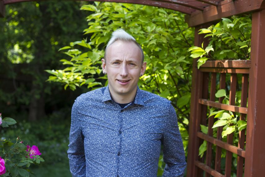 image of Dr. Adam Davies from wasit up. He is wearing a blue shirt and there is a greenery and a garden in the background