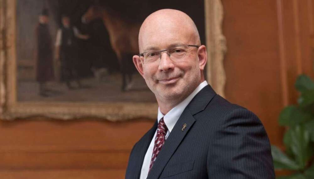 A person in a black suit, white shirt and red tie with wire frame glasses smiles in front of a painting of a horse on a wood panelled wall.