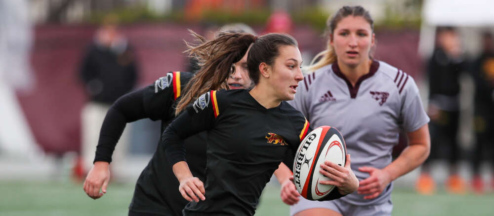 University of Guelph's Talia Hoffman sports a black uniform and holds a white, black, and red rugby ball during a game.