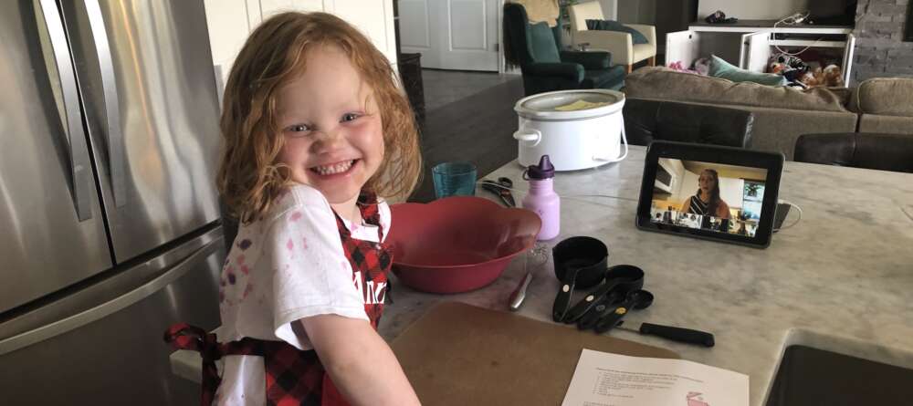 A young girl wearing an apron smiles at the camers while standing at a kitchen counter with a recipe and baking utensils in front of her
