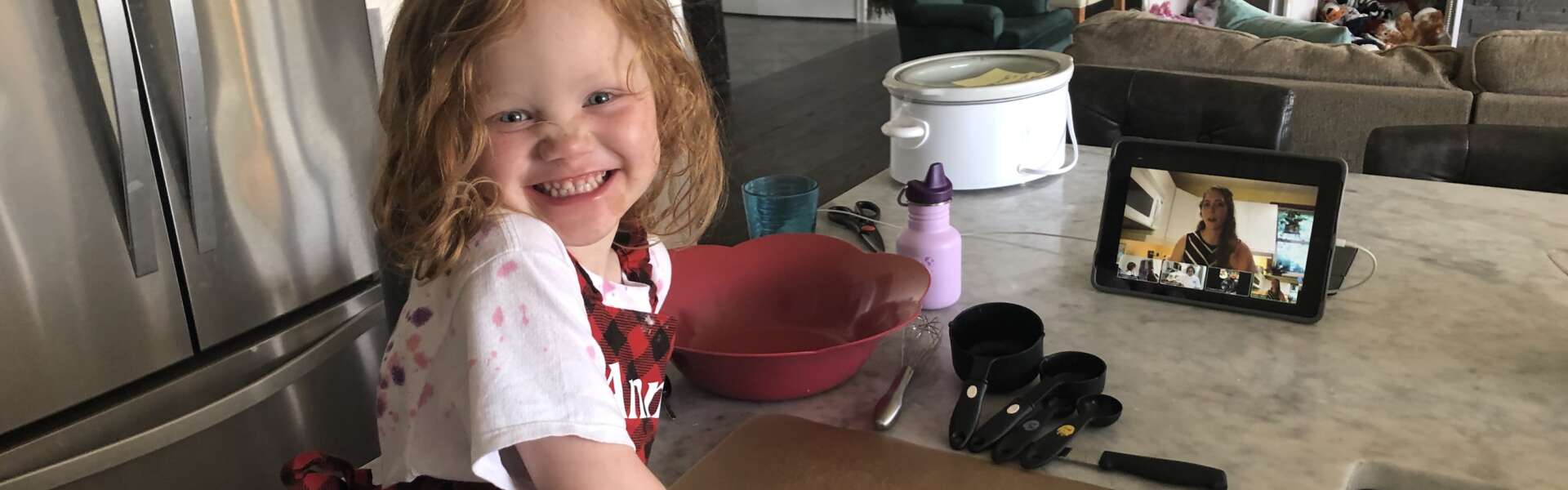 A young girl wearing an apron smiles at the camers while standing at a kitchen counter with a recipe and baking utensils in front of her