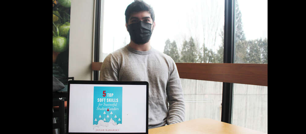 Jonah Kawarsky sits in front of a window. On the table in front of him is his laptop facing the viewer and displaying the cover of his ebook.