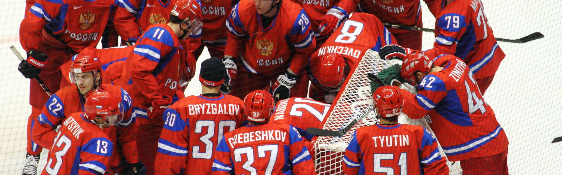 Russian Olympic Hockey players, wearing vibrant red jerseys, huddle around the net