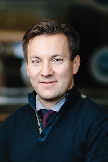 A man with short brown hair faces the camera wearing a shirt and tie with a dark sweater over it.