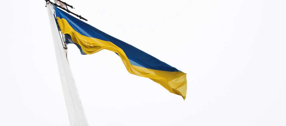 The Ukrainian flag (top half blue, bottom half yellow) flies against a white overcast sky.