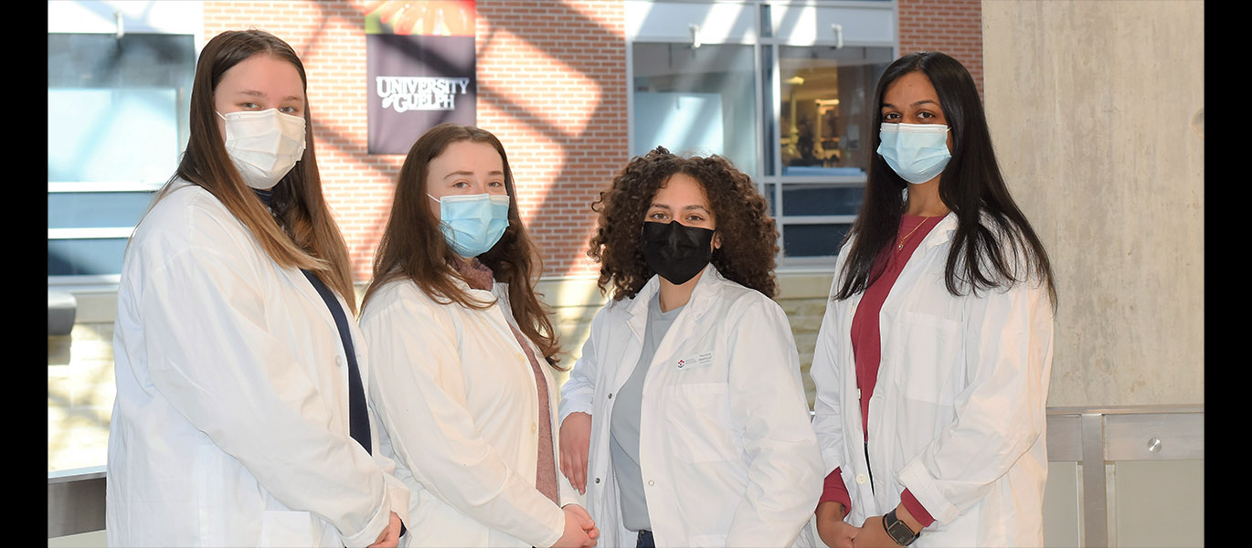 Four women wearing masks and lab coats pose in the Summerlee Science building atrium