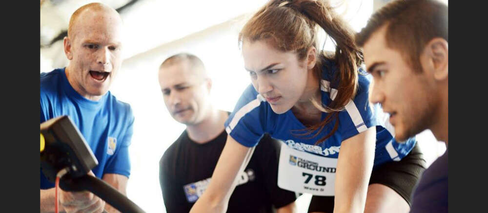athlete Avalon Wasteneys frowns while on a stationary bike surrounded by 3 coaches
