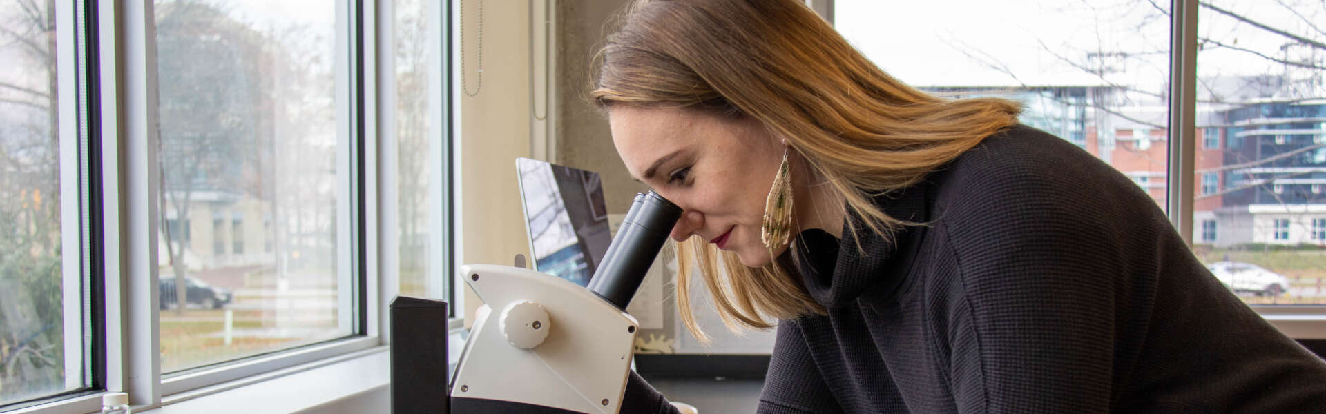 Danielle Nowosad in black sweater leans to peer into a microscope in front of a window.