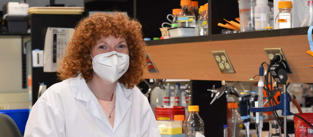 Dr. Jennifer Geddes-McAlister in lab coat sits at station in science lab.