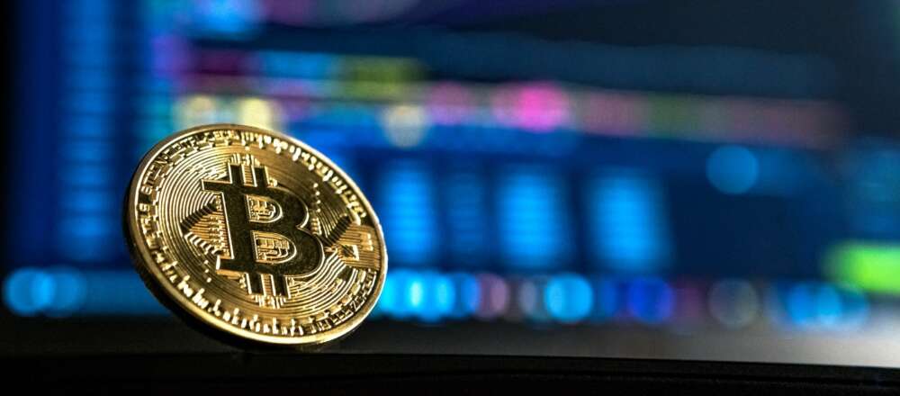 A gold bitcoin, with a capital B, sits in front of a computer monitor displaying stock market trends.