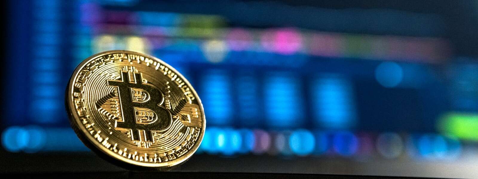 A gold bitcoin, with a capital B, sits in front of a computer monitor displaying stock market trends.