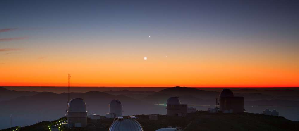 A bright orange sunrise over SO's La Silla Observatory in northern Chile. In the centre of the sky are three shiny lights: Jupiter (top), Venus (lower left), and Mercury (lower right) forming a triangle.