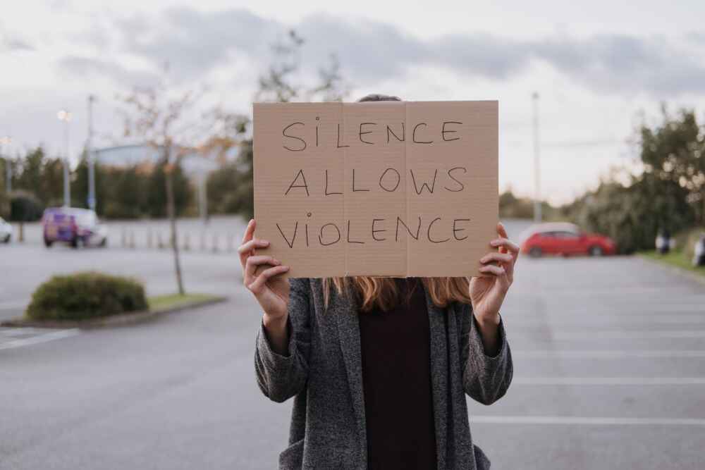 Unrecognizable woman in casual clothes standing on road and demonstrating poster Silence Allows Violence