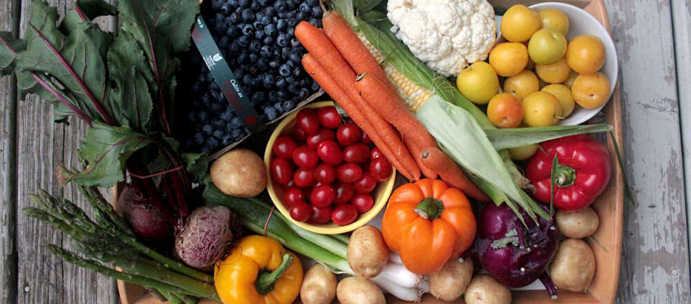 A tray full of fruits and vegetables on a grey, wooden background. On the top part of the tray there is, from left to right, a basket of blueberries, carrots, cauliflower and a bowl of yellow plums. Across the middle of the tray there are beets, potatoes, a bowl of red cherry tomatoes, corn, and a pepper. On the bottom part of the tray there is asparagus, another pepper, potatoes, green onions and kohlrabi.