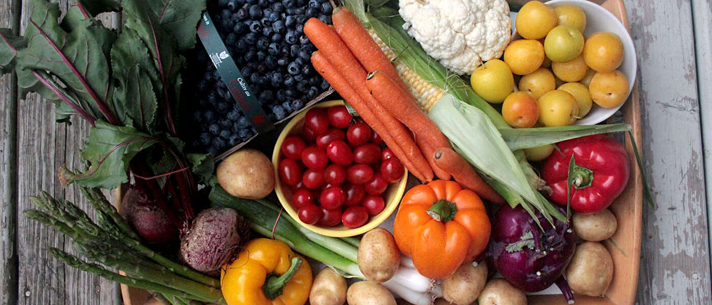 A tray full of fruits and vegetables on a grey, wooden background. On the top part of the tray there is, from left to right, a basket of blueberries, carrots, cauliflower and a bowl of yellow plums. Across the middle of the tray there are beets, potatoes, a bowl of red cherry tomatoes, corn, and a pepper. On the bottom part of the tray there is asparagus, another pepper, potatoes, green onions and kohlrabi.