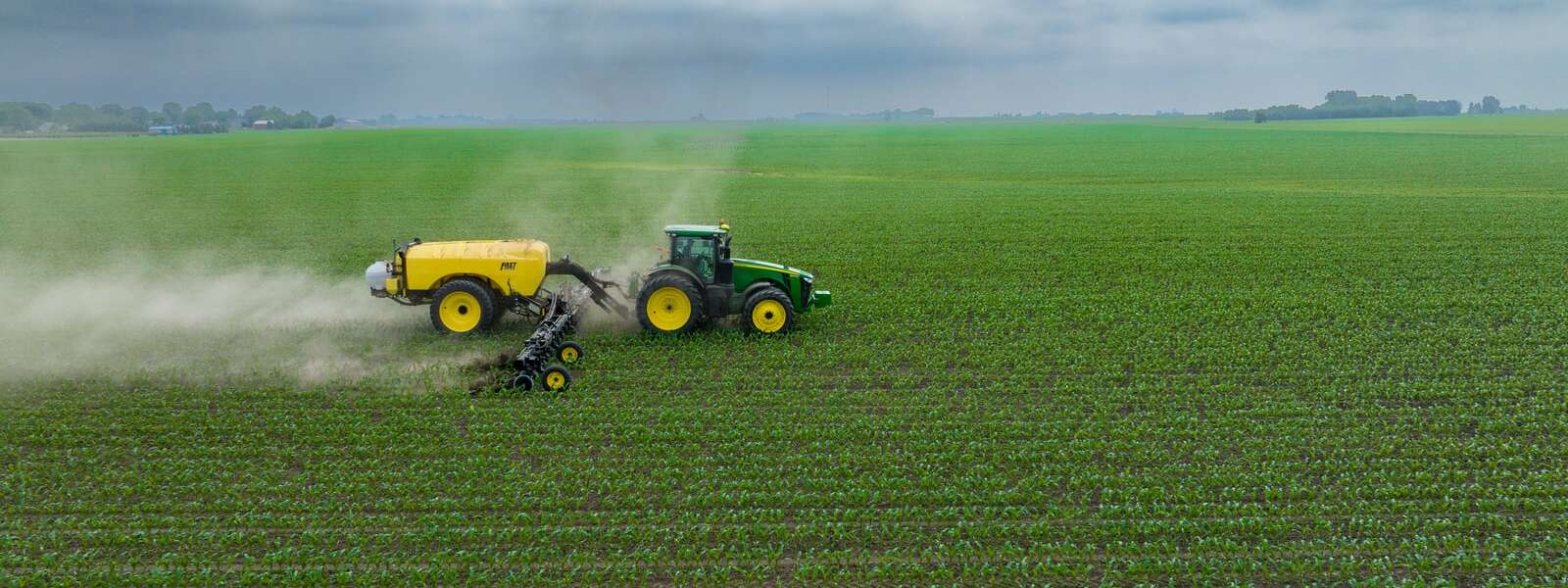 A green tractor with a yellow tank in a green field. The tractor is on the left side of the image and is moving towards the right side. Behind it is a plume of a white substance.