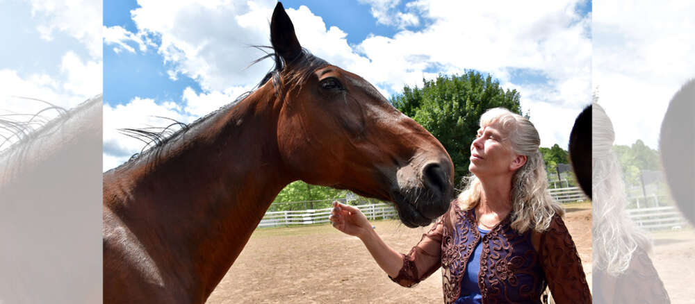 Brown horse stands with Dr. Katrina Merkies in purple shirt against a backdrop of greenery and blue sky