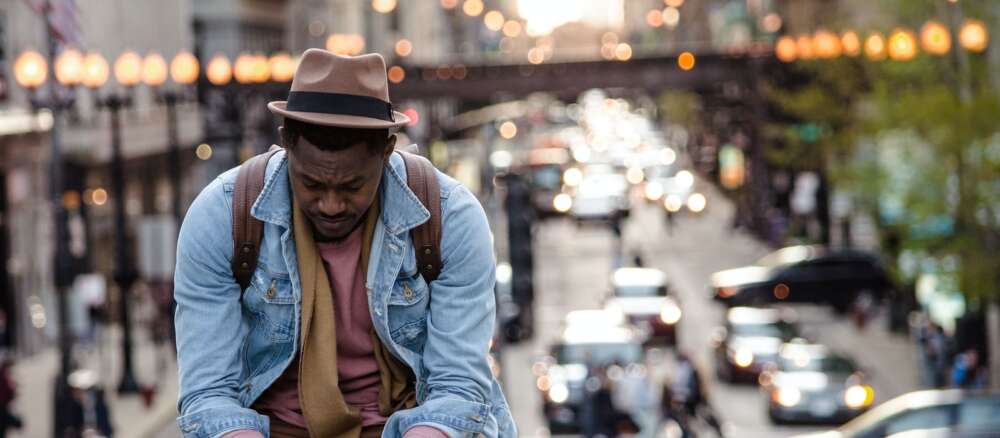 A man in a leather jacket looking down while sitting on a ledge in a city