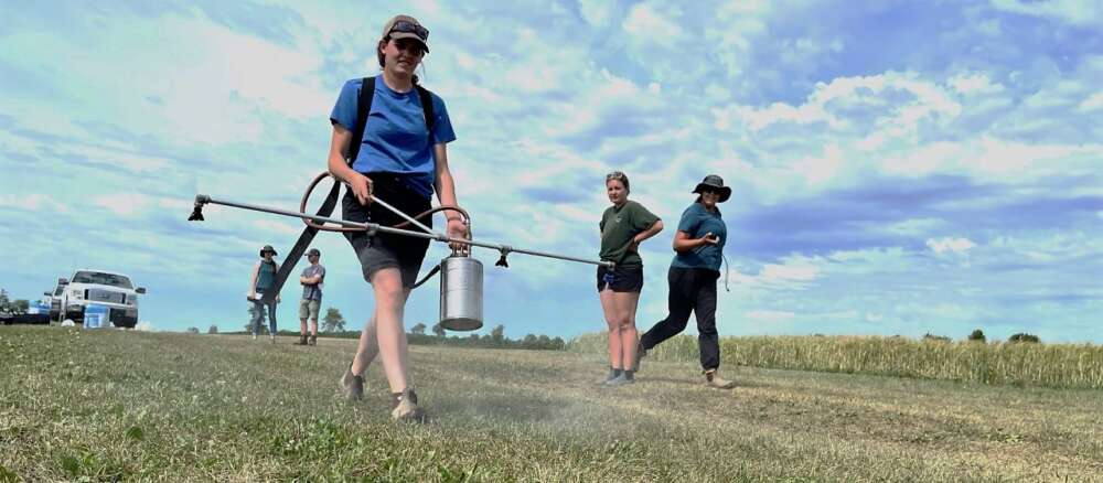 three people walk along a field, one holding a 4-nozzle sprayer bar and canister