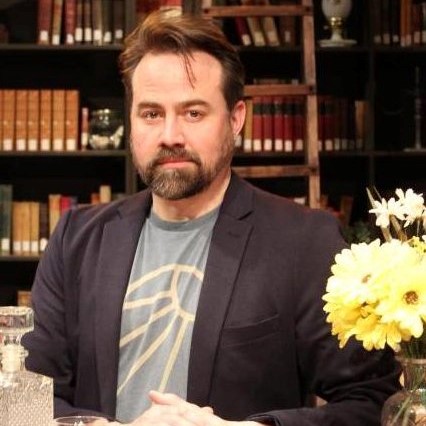 Dr. Kuling sits at a table with yellow flowers and a glass bottle. Behind them is a bookshelf.