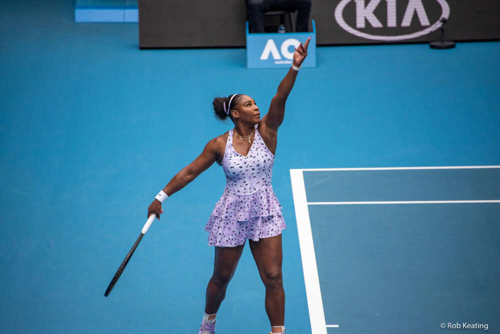 Serena Williams wearing a purple outfit serves on a blue tennis court.