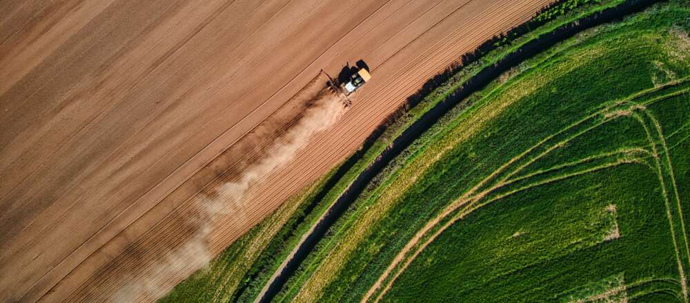 An aerial view of fields, which spilt the image in half diagonally across the centre. In the top half a tractor drives through a brown field. In the bottom half is a green field.