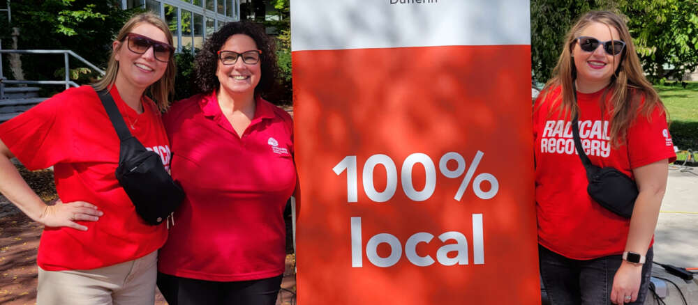 Three people wearing red shirts stand next to a sign reading "100% local"