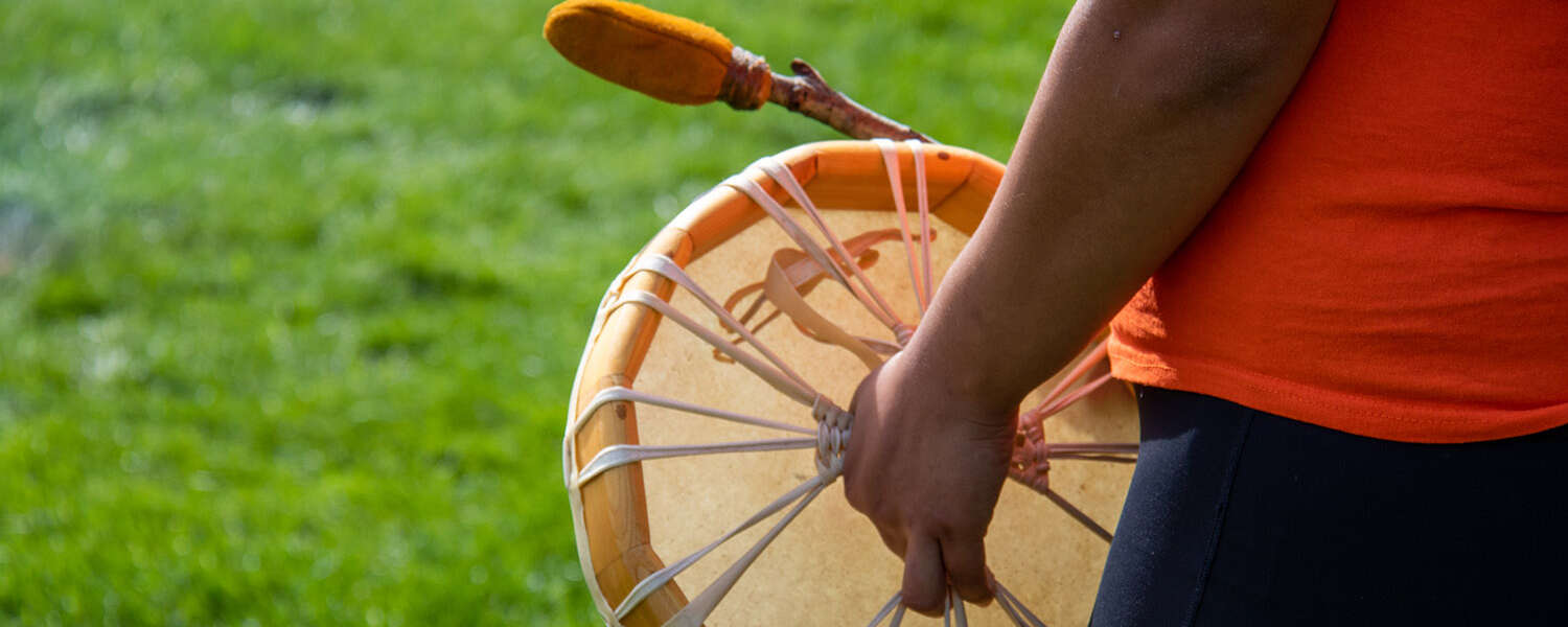 A closeup of the hand of a person wearing an orange shirt and holding a traditional drum