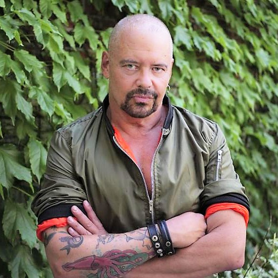 Prof. Sky Gilbert crosses his arms and stands against an ivy-covered wall