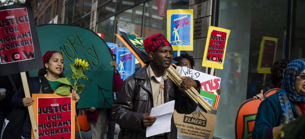 A man in brown clothes, leather jacket and red head scarf carries protest sign with crowd of people.