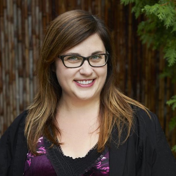 Dr. Sara Epp poses for a headshot in front of a wooden wall next to cedar trees.