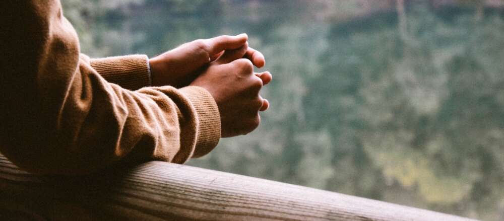A person wearing a tan jacket, with only their arms and hands visible. leans against a railing overlooking trees and a lake.