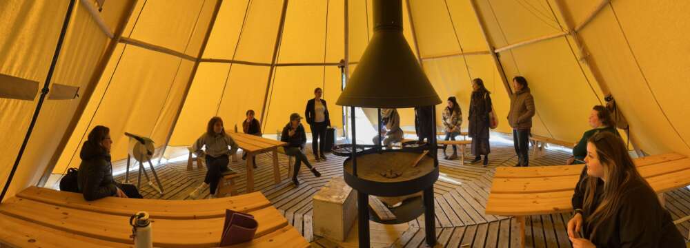 People sit on wooden benches inside a circular tent with a woodstove in the middle.