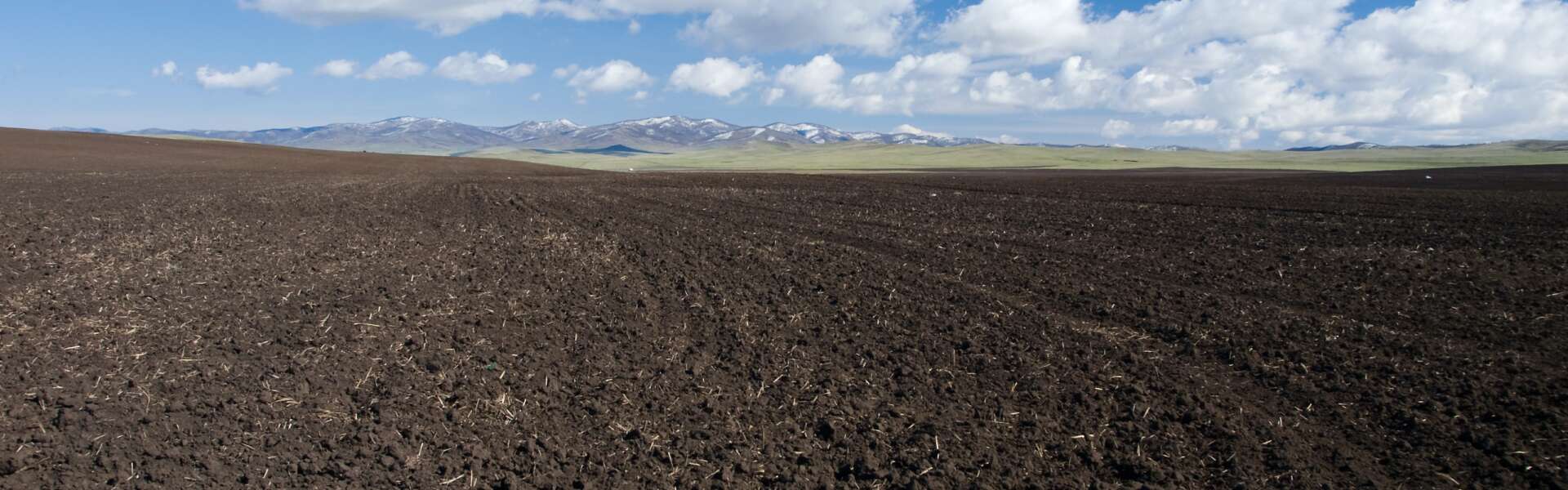 A tilled field with mountains in the distance and a cloudy blue sky.