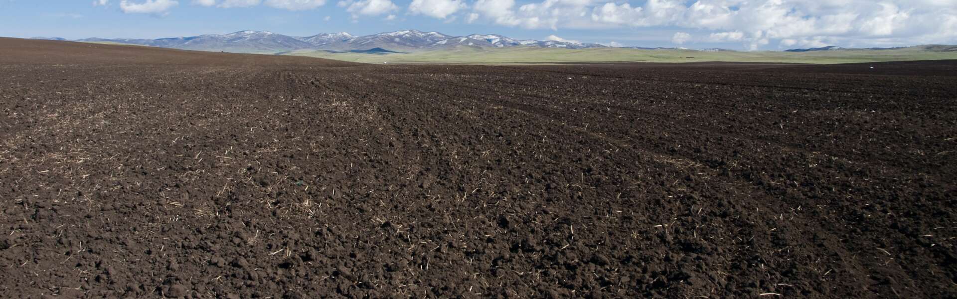A tilled field with mountains in the distance and a cloudy blue sky.