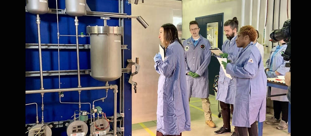 Four people in lab coats look inside a large blue chamber featuring water filtration pipes on the side