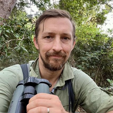 Dr. Travis Steffens takes a selfie while holding a pair of binoculars in a forest